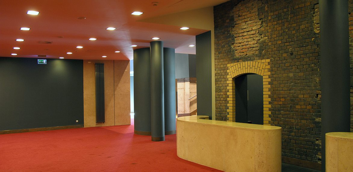 Wnętrze korytarz teatru. Czerwony kolor sufitu i podłogi, filary w kolorze szarym.